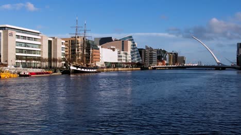 Eine-Langsame-Zoom-aufnahme-über-Wasser-Des-Flusses-Liffey-In-Der-Abendsonne,-Die-Den-North-Wall-Kai-Mit-Der-Samuel-Beckett-Bridge-In-Der-Ferne-Zeigt