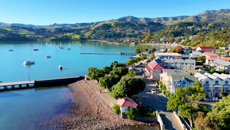 aerial footage captures akaroa's vibrant harbor, lush hills, and charming town under bright daylight, highlighting the serene coastal landscape