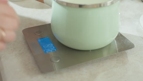 close up of modern mint green pot being placed on digital kitchen scale by person as soft shadow reflects nearby while preparing to pour ingredients into pot on clean countertop