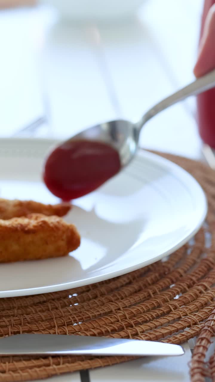 Un cucchiaio versa il ketchup sul pollo fritto su un piatto bianco, posizionato su una tovaglietta intrecciata con illuminazione brillante