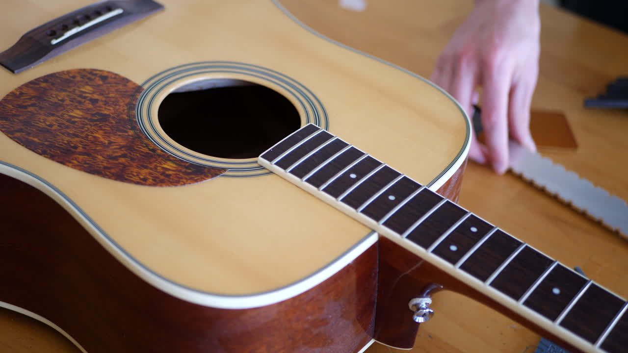 Close up on the hands of a luthier craftsman leveling an acoustic guitar neck and fretboard while adjusting the trussrod on a workshop bench with lutherie tools