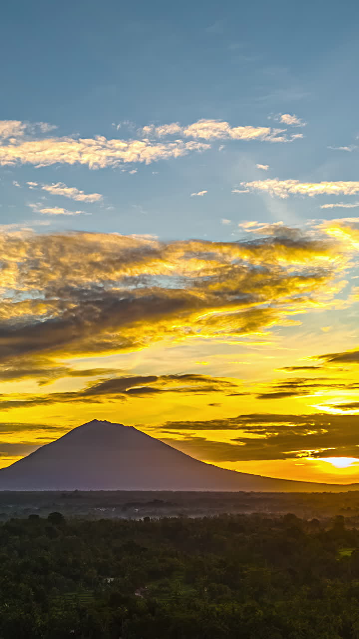 Mount Agung silhouette at sunrise with golden sky over forest in Bali, vertical timelapse