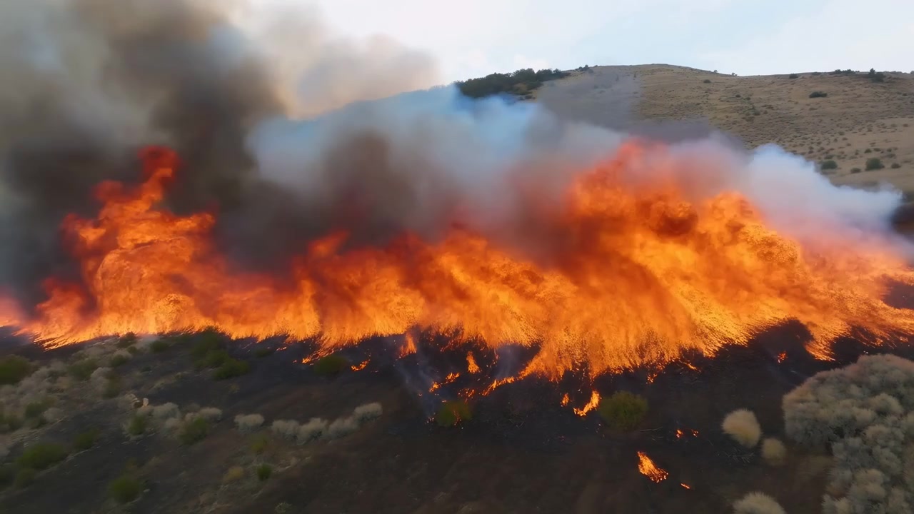 Fiamme vivaci avanzano attraverso la bassa vegetazione su un terreno arido e ondulato, mentre un fumo denso si alza in nuvole stratificate grigie, marroni e bianche sopra terra bruciata e arbusti sparsi
