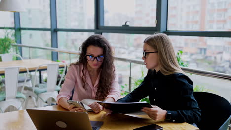 mid-aged young business ladies discussing job issues. long-haired women in glasses sitting at desk looking at phone and communicating.