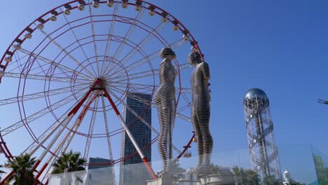 ferris wheel against the blue sky with statue of ali and nino on embankment of batumi