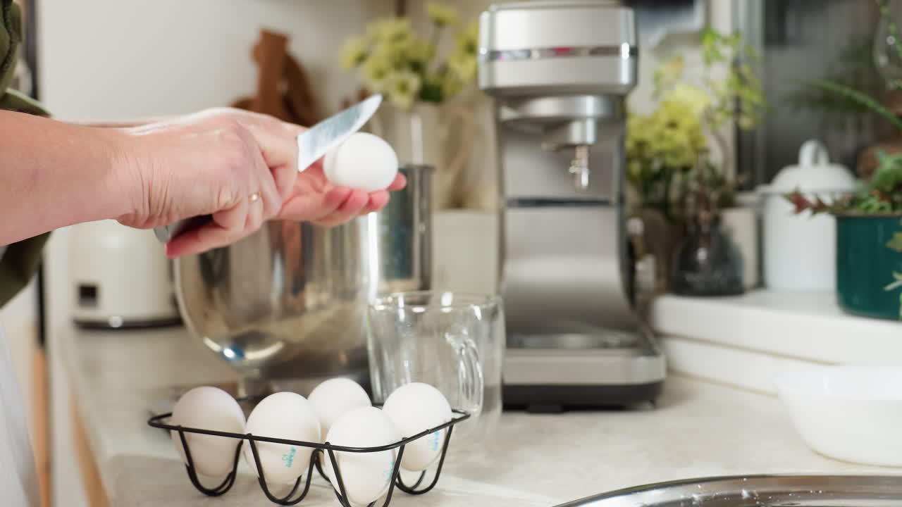 Close up of person in kitchen taking egg from black wire rack, cracking it gently with knife, pouring egg white into stainless bowl while carefully preventing yolk from dropping