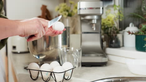 close up of person in kitchen taking egg from black wire rack, cracking it gently with knife, pouring egg white into stainless bowl while carefully preventing yolk from dropping