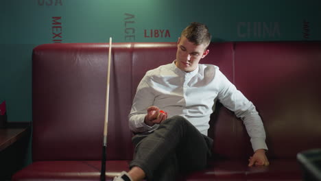un joven con camisa blanca y pantalones a cuadros se sienta en el sofá rojo en la sala de billar, rodando una pelota de billar naranja en la mano. el palo de la señal se inclina a su lado. el fondo presenta nombres de países en la pared verde