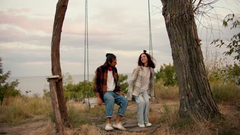 Shooting-from-behind:-A-guy-and-his-girlfriend-in-checkered-shirts-sit-on-a-wooden-swing-and-start-swinging.-Rest-in-the-country-by-the-sea