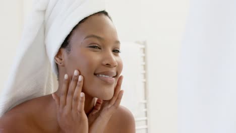 happy biracial woman with towel on head looking in mirror touching her face in bathroom, slow motion