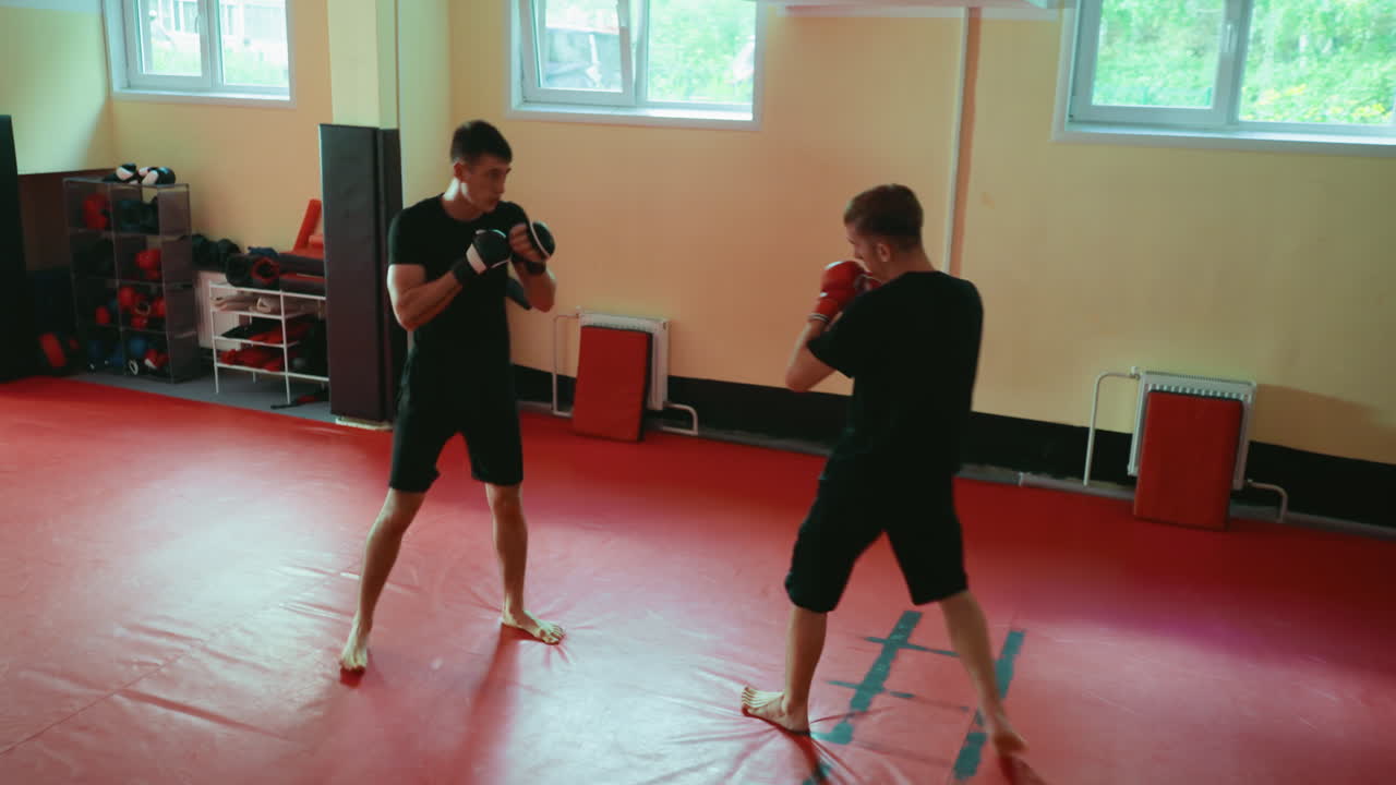 Two boxers sparring in martial arts gym throwing punches while opponent defends with guard on red mat floor during training session under natural window light with equipment