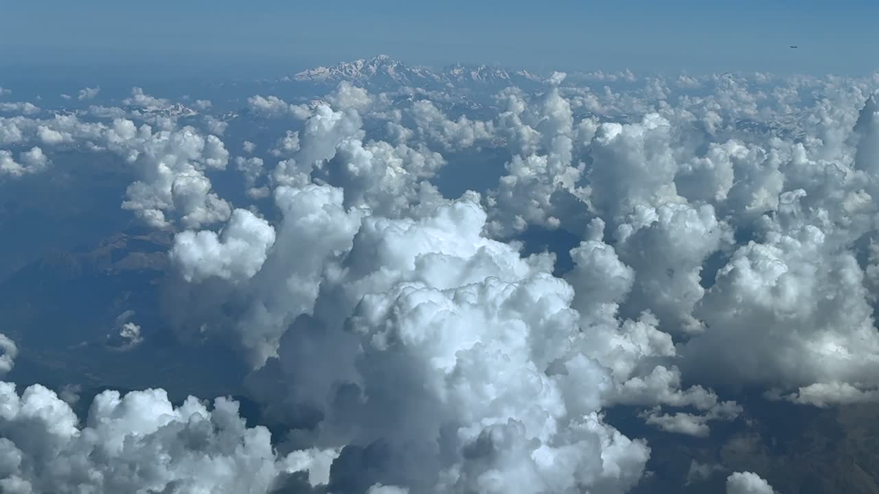 An elevated aerial view from a jet cockpit while flying over cottony cumulus clouds over the Alps mountains, with the Mont Blanc peak in the distance. A cockpit view in a hazy summer morning.