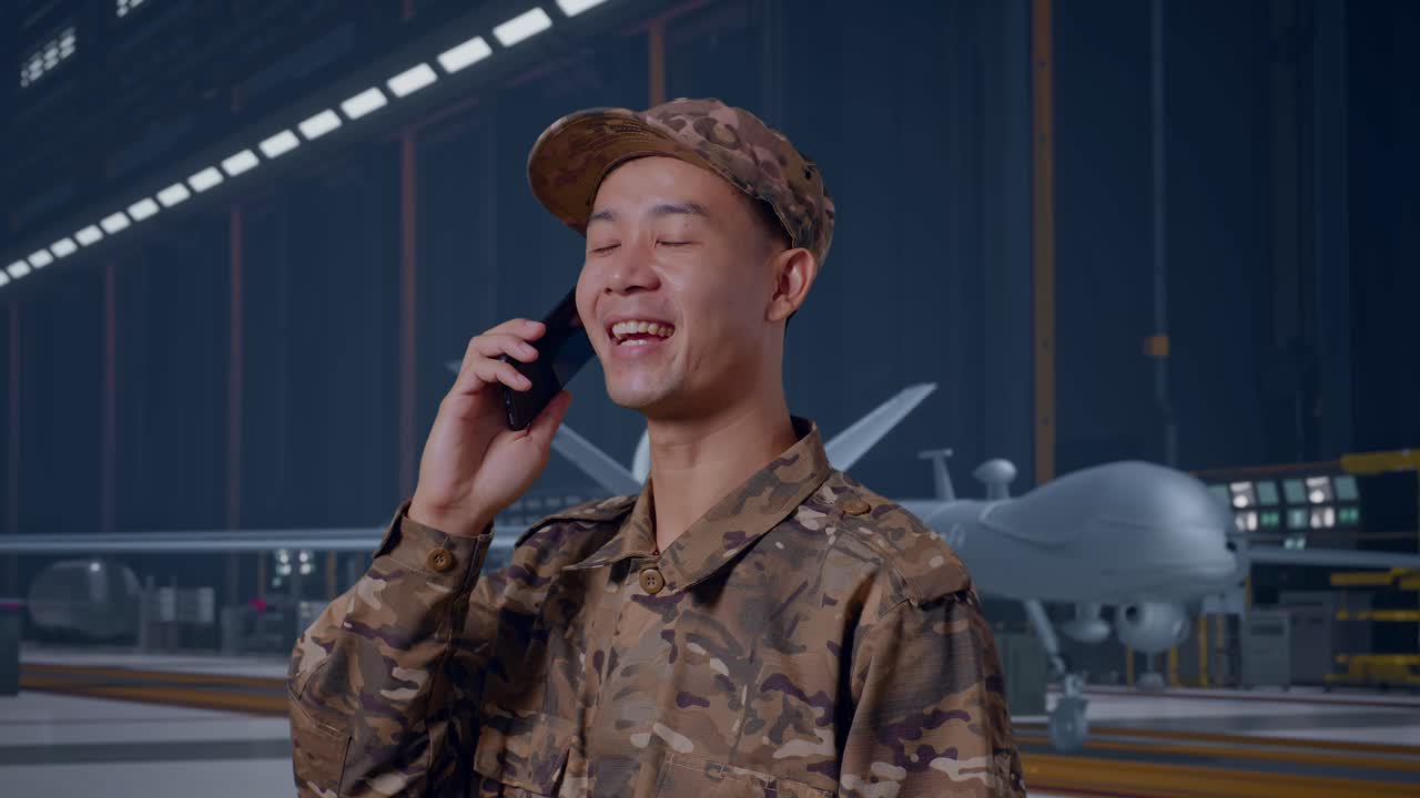 Primo piano laterale di un soldato asiatico che parla allo smartphone e si guarda intorno, mentre si trova accanto a un moderno drone militare in un hangar di manutenzione