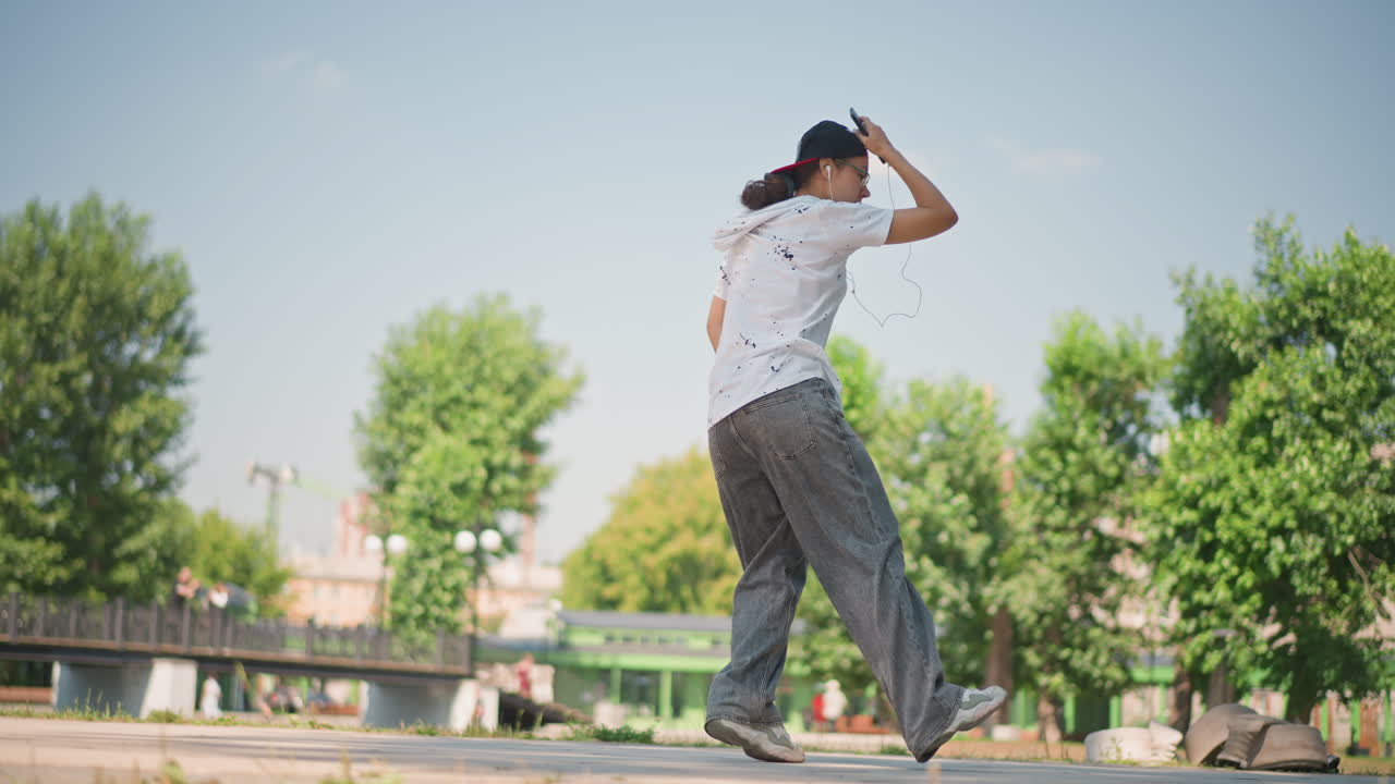 햇빛이 잘 드는 광장에서 자유형을 선보이는 사람이 균형 잡힌 자세로 포즈를 취하고 하이킥을 하며, 나뭇잎이 무성한 나무가 있는 탁 트인 공원 배경, 자신감 넘치는 자세와 표현력이 풍부한 리허설 분위기를 연출해요.