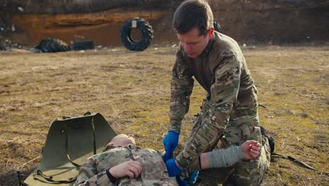 A-confident-man-in-camouflage-clothing-with-blue-elastic-medical-gloves-binds-the-hand-of-an-injured-man-with-a-tourniquet-during-combat-operations-in-the-steppe.-Providing-personal-assistance-to-wounded-soldiers-during-combat-operations