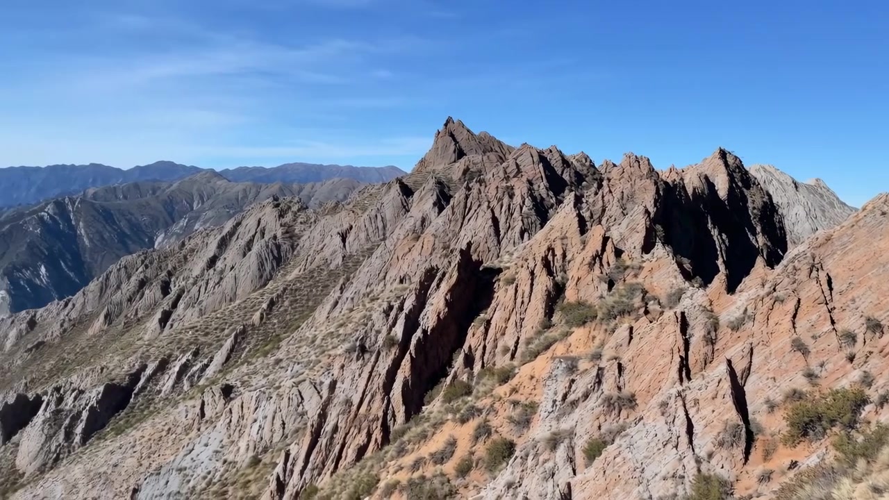Formazioni rocciose montuose rimangono in vista mentre il movimento della telecamera rivela creste stratificate, scogliere rocciose, piante sparse e una luce diurna intensa attraverso un vasto terreno naturale con cime distanti.