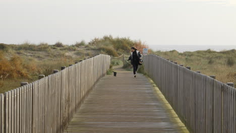 Middle-age-woman-with-miniature-dachshund-dog-running-together-slow-motion-across-wooden-beach-boardwalk