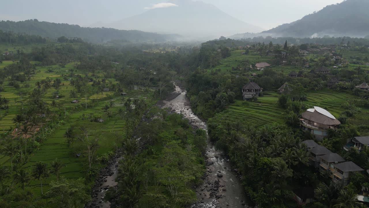 un río rodeado de exuberantes campos de arroz verdes que conducen a una vista nebulosa del volcán del monte agung, bali