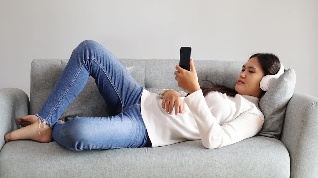 Giovane donna asiatica che usa lo smartphone e scorre i social media mentre è sdraiata sul divano a casa e si rilassa.
