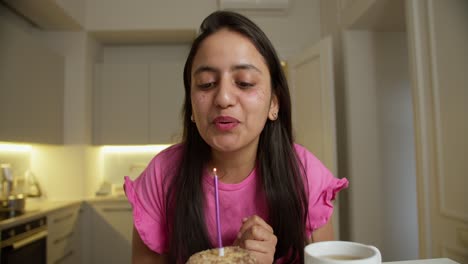 happy brunette girl in a pink dress claps her hands and blows out a pink candle on her small cake while sitting at a table in a modern apartment