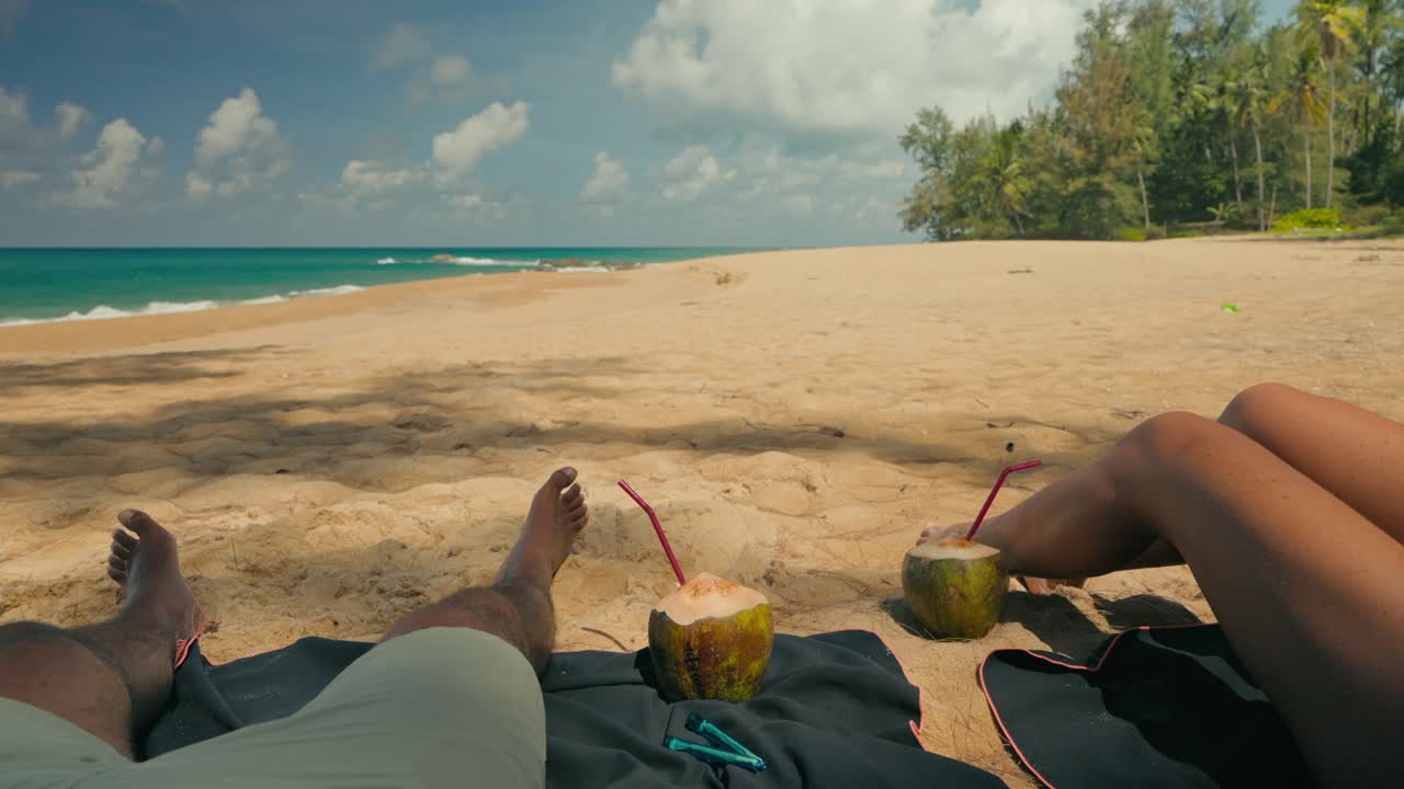 Primo piano delle gambe di una coppia che si rilassa su una spiaggia tropicale, con noci di cocco e due sigarette arrotolate nelle vicinanze. Onde soleggiate, sabbia e palme creano un'atmosfera di vacanza tranquilla e rilassata.