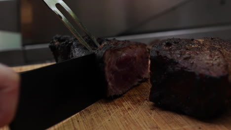 Thick-juicy-cooked-steak-sliced-with-fork-and-large-knife-on-wood-chopping-board,-close-up-side-view