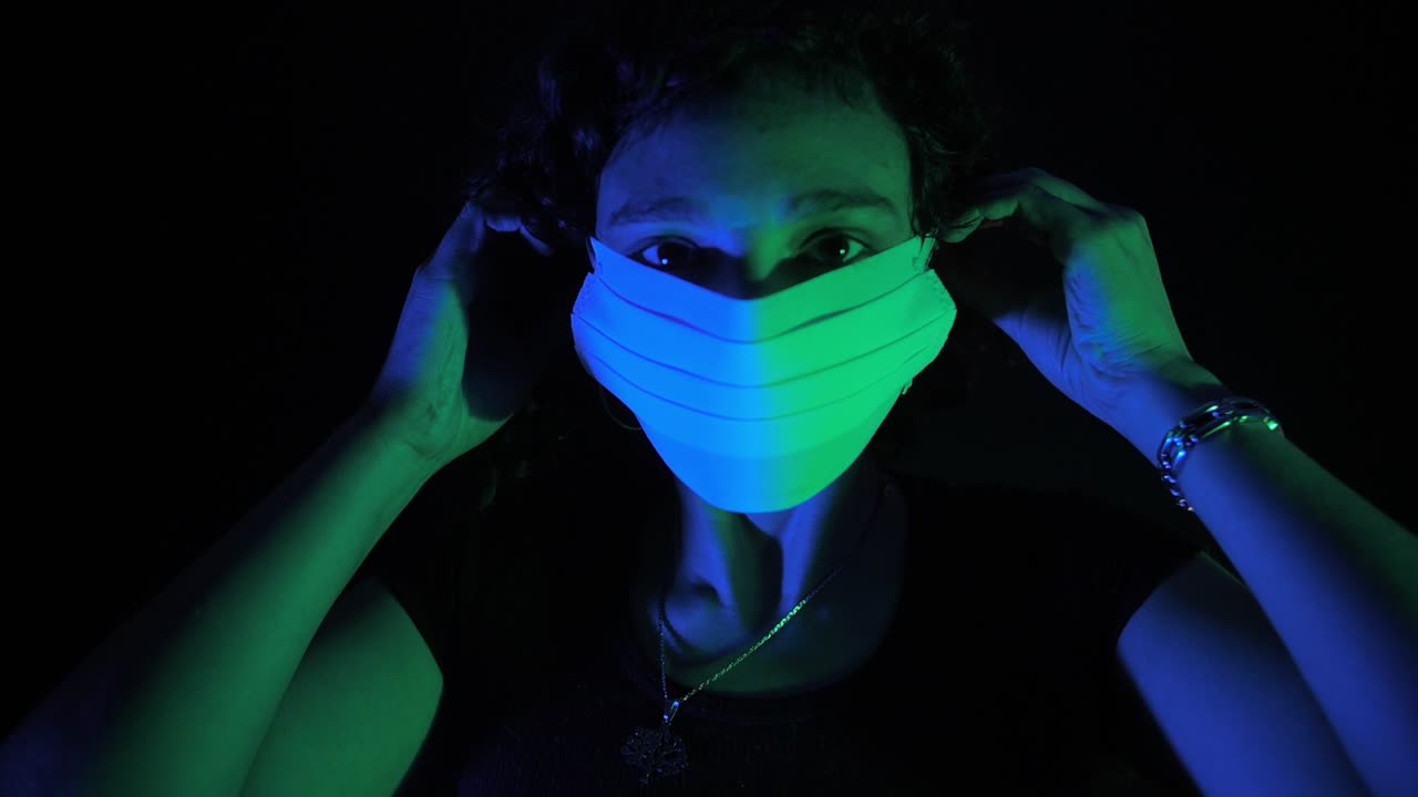 Jovem mulher coloca máscara facial médica com iluminação dramática azul e verde.