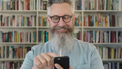 portrait of mature caucasian professor with beard texting browsing using smartphone researching information in library bookshelf background