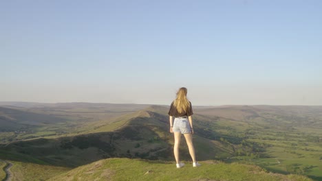 stabilised shot of young blonde woman standing on top of mam tor, castleton, peak district, england admiring the view of green rolling hills and blue skies