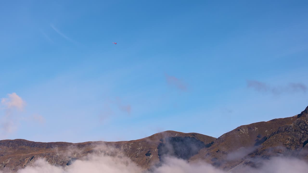 A helicopter flies over misty mountains and Lake Wakatipu under a clear blue sky
