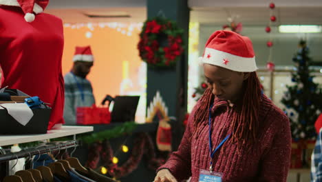 Happy-retail-assistant-wearing-Santa-hat-walking-through-clothing-rack-rows-in-Christmas-decorated-shopping-mall-store,-using-clipboard-to-crosscheck-price-tags,-making-sure-shop-is-ready-for-clients