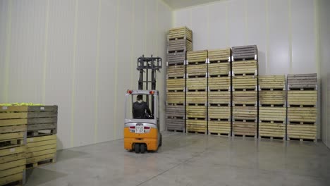 A warehouse worker carries wooden boxes of apples and stacks them in rows together near the wall of the room. Juice factory.