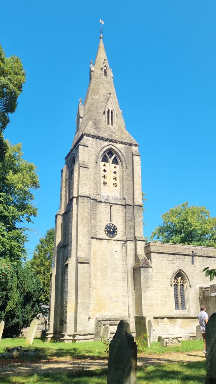 Vertical shot of St Mary's Church in Bainton, Cambridgeshire, UK, with a male visitor strolling past under a bright blue afternoon sky, capturing the historic charm of the religious building