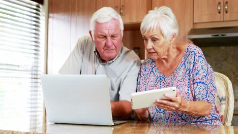 una pareja de ancianos usando una computadora portátil y una tableta digital en la cocina