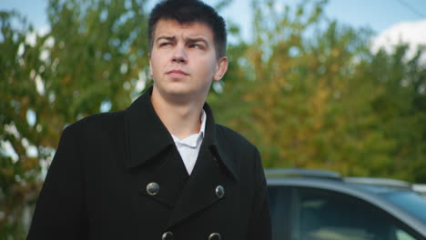 investor near car adjusting collar with both hands outdoors in professional black coat, confidently preparing for business under clear blue sky with green foliage and vehicles in urban background