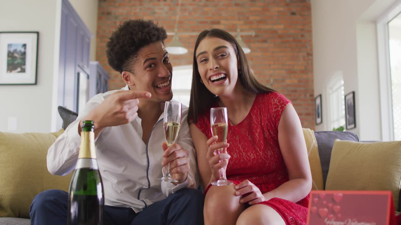 feliz pareja biracial bebiendo champán en casa haciendo una celebración llamada de video portátil y riendo