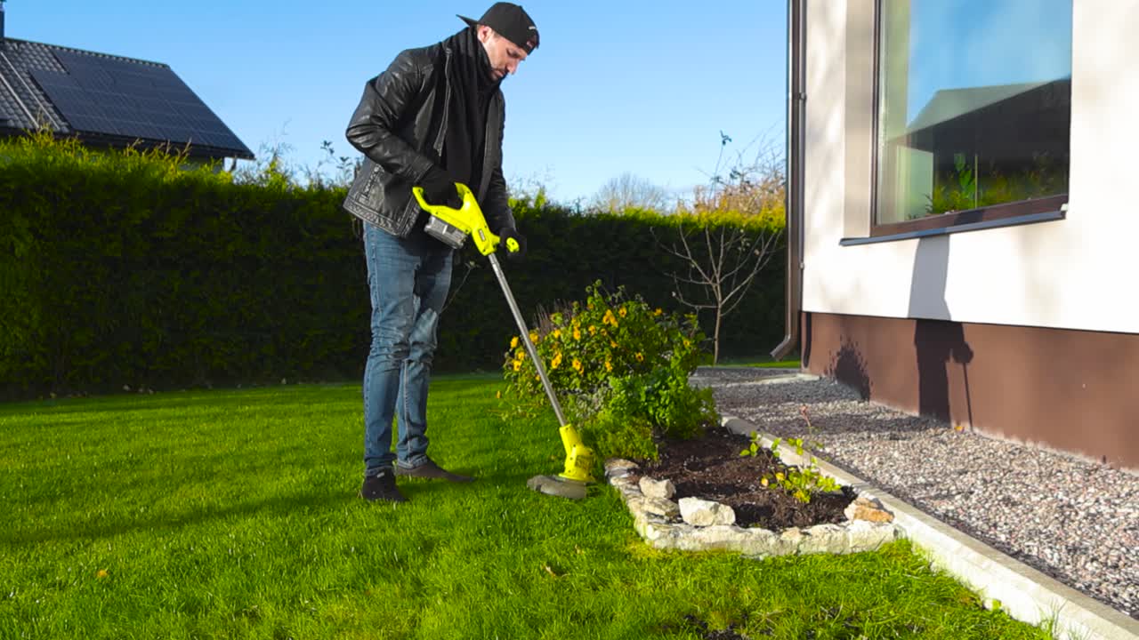 Man working at a sunny autumn garden with a electric lawn trimmer or grass trimmer, working on limestone garden bed edge. Grass is flying in slow motion and the man is creating shadows on house wall.