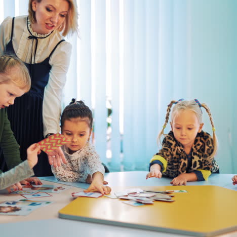 little students learning english with pictures at school. teacher controls educational process of children who sit at round table and look at cards in the light classroom.