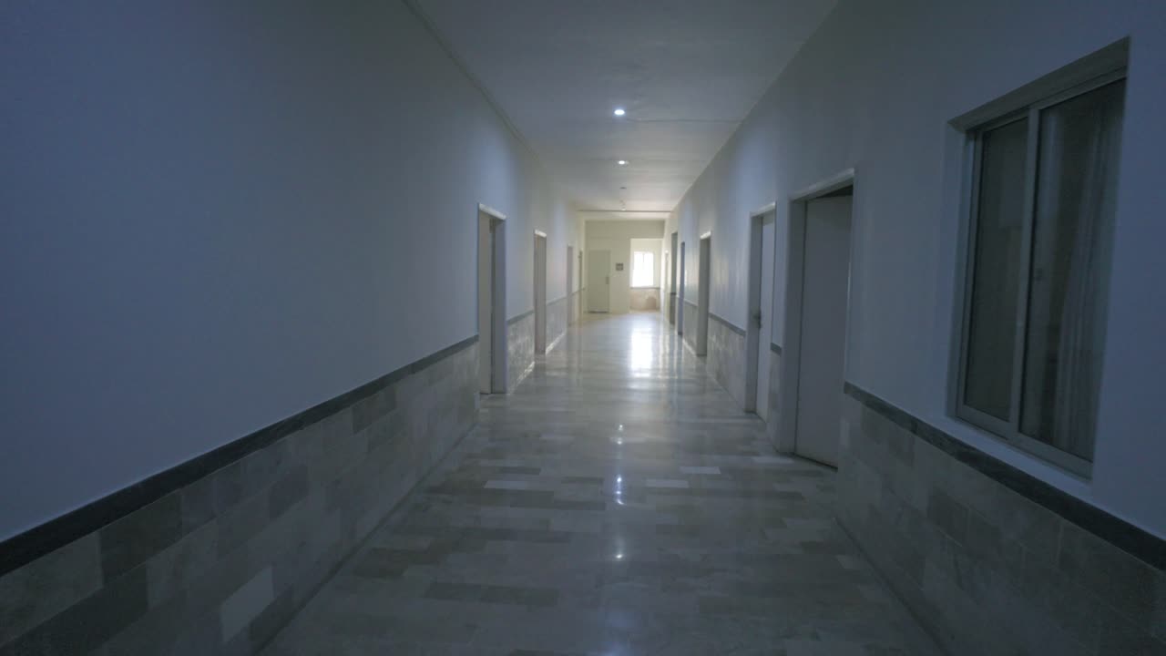 A quiet hospital corridor with reflective marble flooring, open side doors, and distant daylight filtering through a rear window, captured in a smooth camera motion