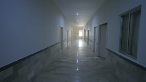 a quiet hospital corridor with reflective marble flooring, open side doors, and distant daylight filtering through a rear window, captured in a smooth camera motion