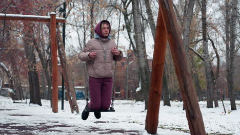 dama vestida cálidamente para el invierno en el columpio en el parque sosteniendo cadenas, con pantalones de color marrón y chaqueta de invierno, con nieve en el suelo y árboles en el fondo