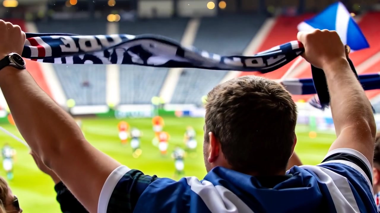 Tijdens lopende wedstrijd draagt fan blauw shirt, houdt teamscarf vast op tribune, zwaait met Andreaskruisvlaggen en juicht.