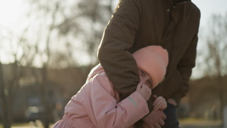 a little girl in a pink cap and jacket playfully hugging and hiding behind a man wearing a brown jacket and blue jeans