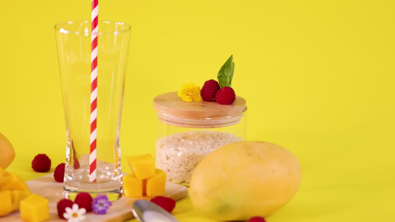 Fresh mango, raspberries, rice, and glass with straw arranged for smoothie preparation, bright lighting