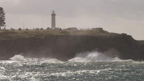 Enfoque-A-Través-De-La-Lluvia-De-Olas-Rompiendo-En-Las-Rocas-Al-Pie-De-Un-Faro