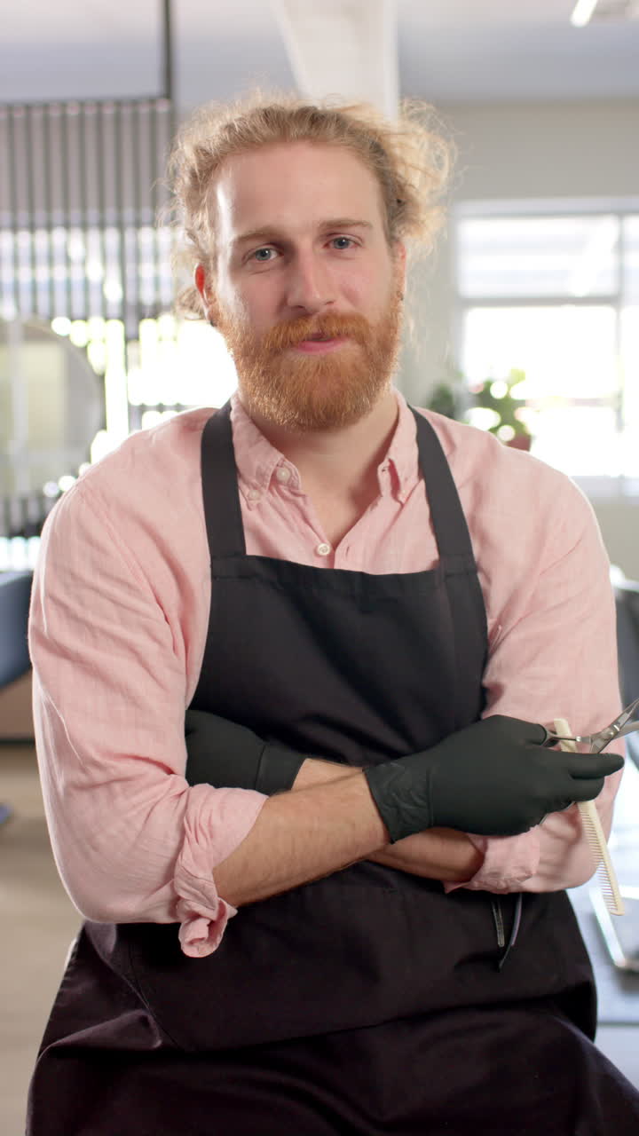 Vertical video of portait of caucasian male hairdresser smiling at hair salon, in slow motion