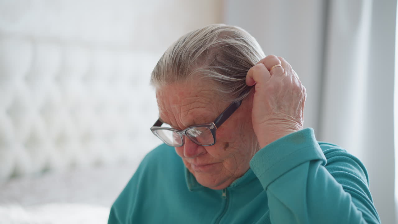 Close-up shot of elderly woman adjusting glasses while gently scratching her head, in soft lighting. Indoor setting with visible wrinkles and age, showcasing a thoughtful or puzzled moment