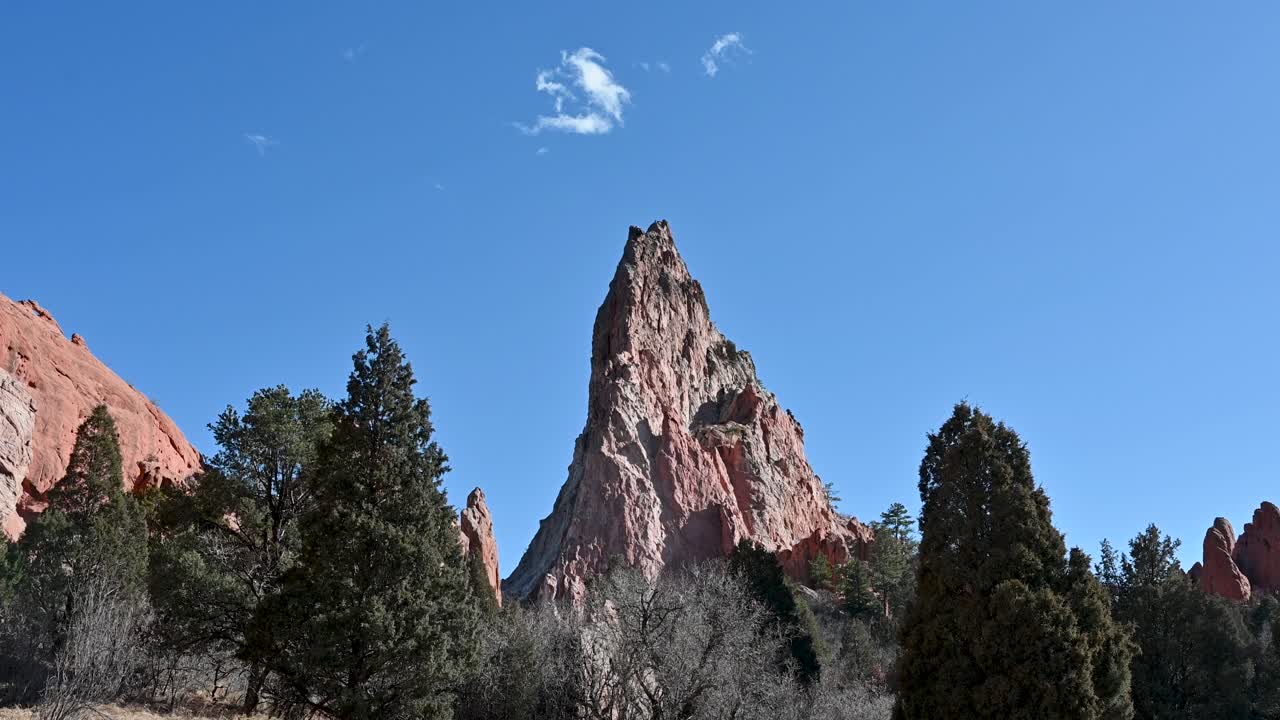 Drone shot of towering red sandstone spire surrounded by pine trees beneath a blue sky at Garden of the Gods in Colorado Springs