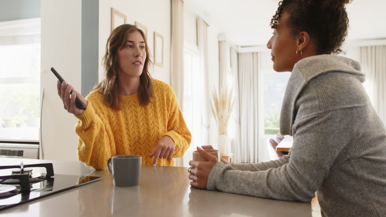 Sharing ideas after remote lift, diverse female friends sipping coffee gesturing at kitchen island