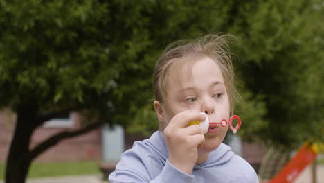 Nahaufnahme-Eines-Kleinen-Mädchens-Mit-Down-Syndrom,-Das-An-Einem-Windigen-Tag-Seifenblasen-Im-Park-Bläst